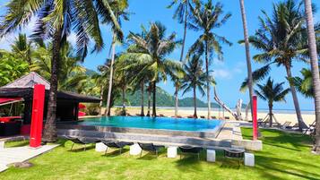 A pool area with lounge chairs and a small structure, surrounded by palm trees and a beach view.