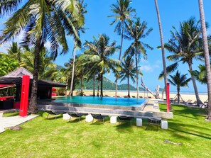 A pool area with lounge chairs and a small structure, surrounded by palm trees and a beach view.