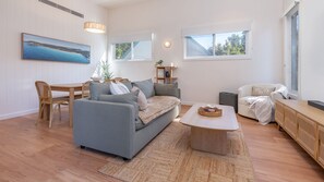 A modern living room with a grey sofa, wooden dining table, and a coffee table. - Schuey's Shack | Luxury | Walk to Boomerang Beach (Boomerang Beach)