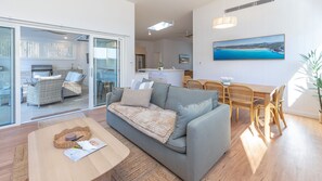 A modern living room with a sofa, a dining table with chairs, a kitchen in the background, and a large window with a view. - Schuey's Shack | Luxury | Walk to Boomerang Beach (Boomerang Beach)