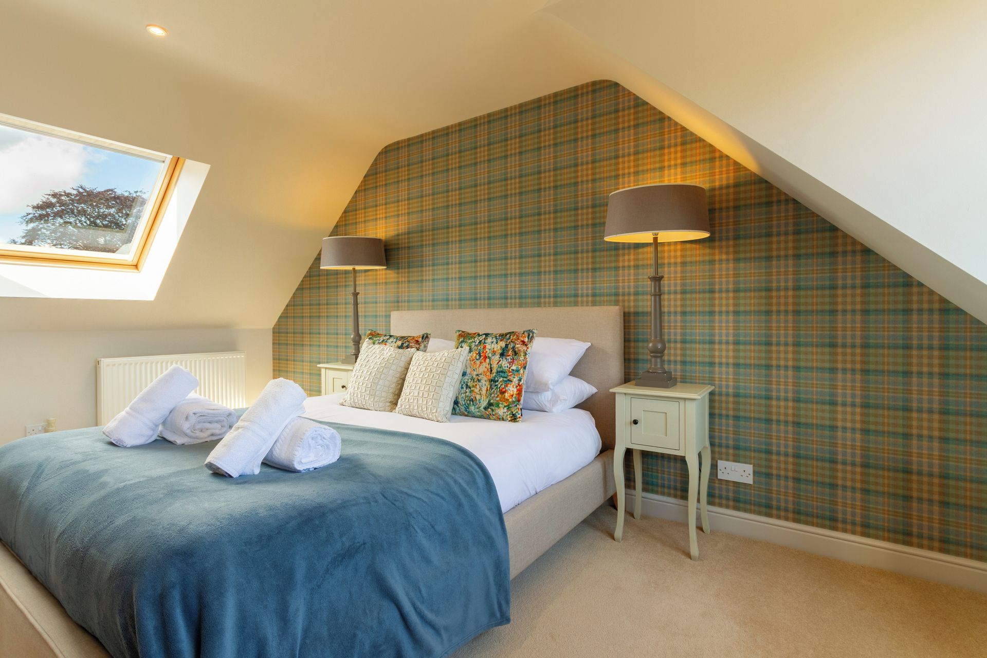 A bedroom with a sloped ceiling, a bed with a blue bedspread, two bedside lamps, and a plaid wallpaper.