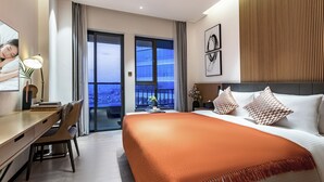Hotelkamer met een bed, een bureau, een stoel, een bureaulamp, een vaas met bloemen en uitzicht op de stad vanuit het raam.