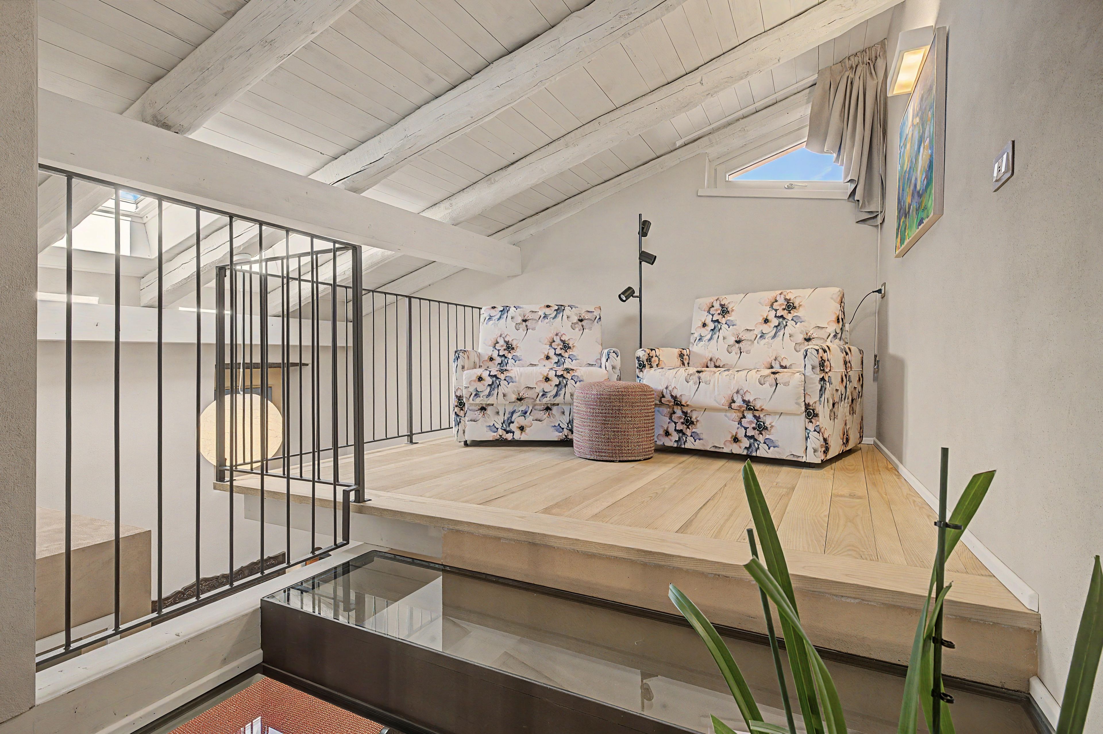 Um quarto amplo no estilo loft, com teto alto, escada de vidro, sofá estampado com flores e uma janela pequena.