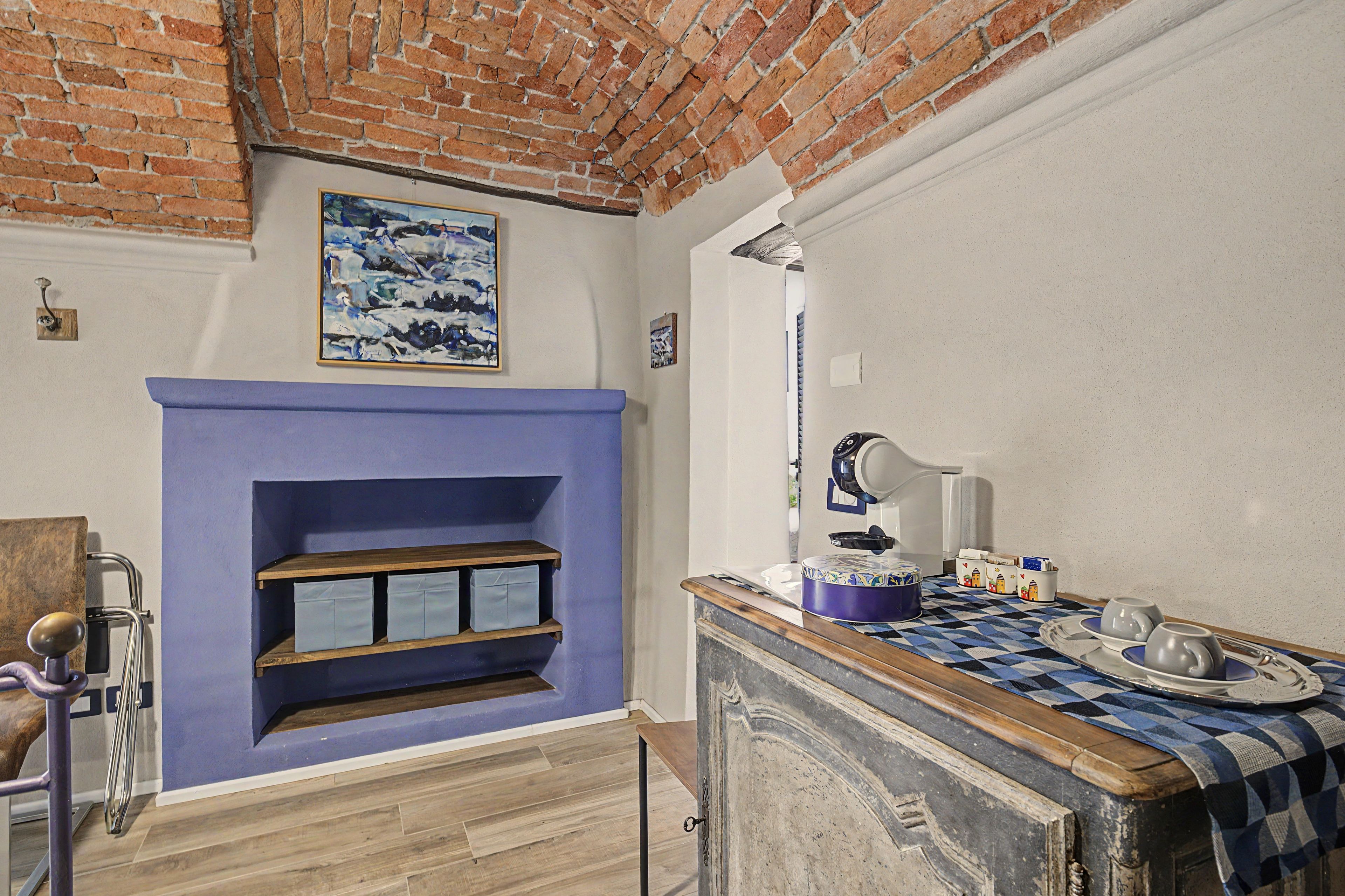 A room with a brick wall, a blue fireplace, a wooden shelf, a table with a checkered tablecloth, and a white lamp.