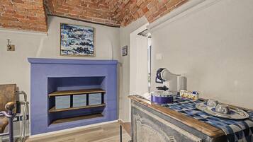 A room with a brick wall, a blue fireplace, a wooden shelf, a table with a checkered tablecloth, and a white lamp.