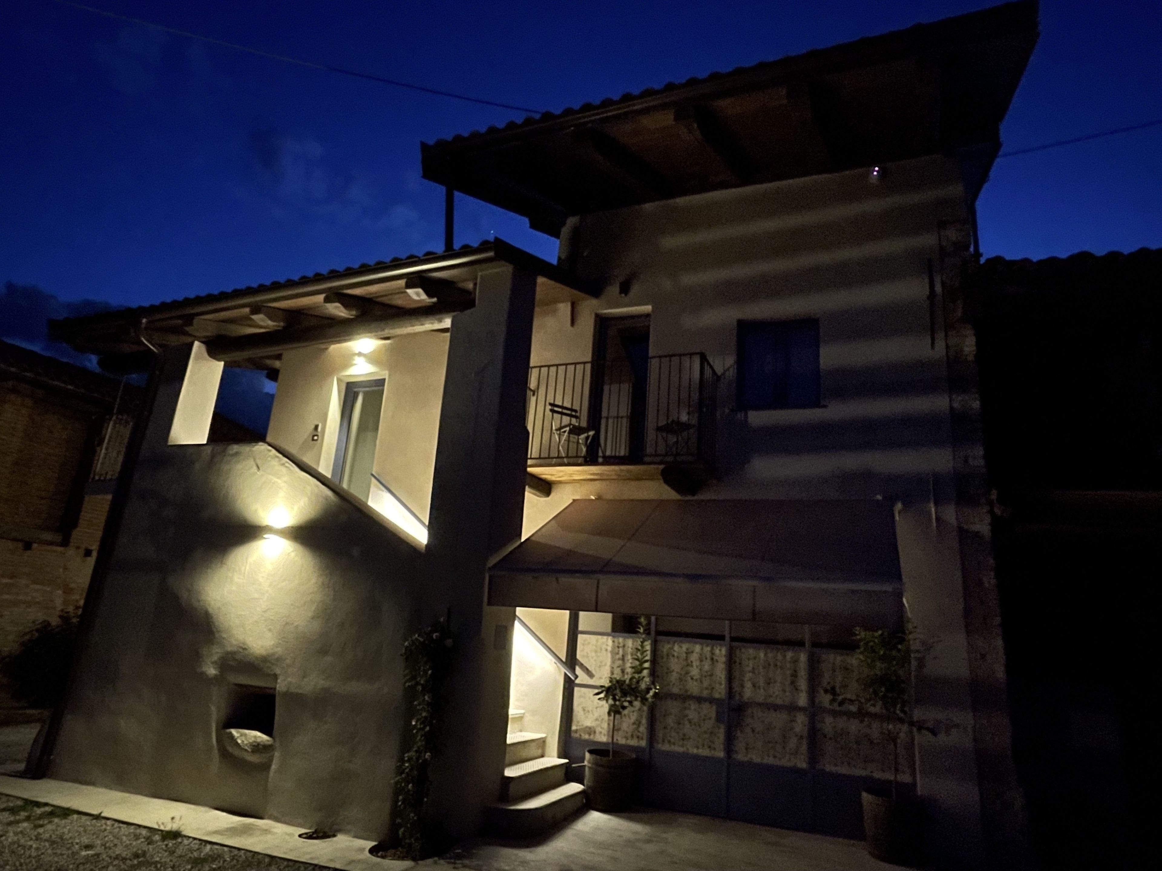Un edificio de dos pisos con un balcón en el nivel superior, una pequeña entrada con escalones, y una fuente de luz que ilumina la zona de acceso.