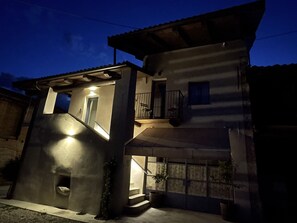 Un edificio de dos pisos con un balcón en el nivel superior, una pequeña entrada con escalones, y una fuente de luz que ilumina la zona de acceso.