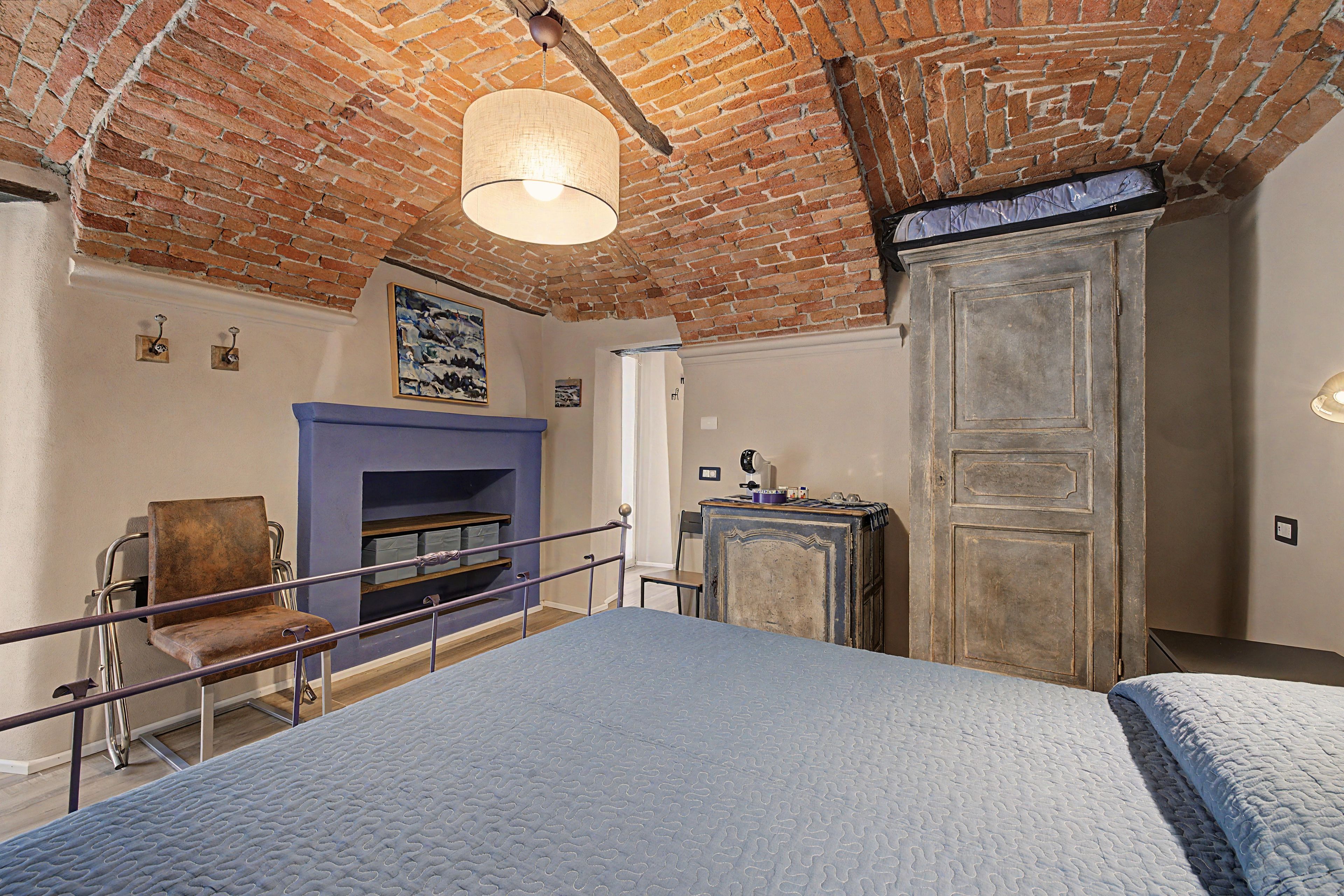 Una stanza con soffitto in mattoni, un letto con materasso blu, una sedia, un tavolino e un armadietto.