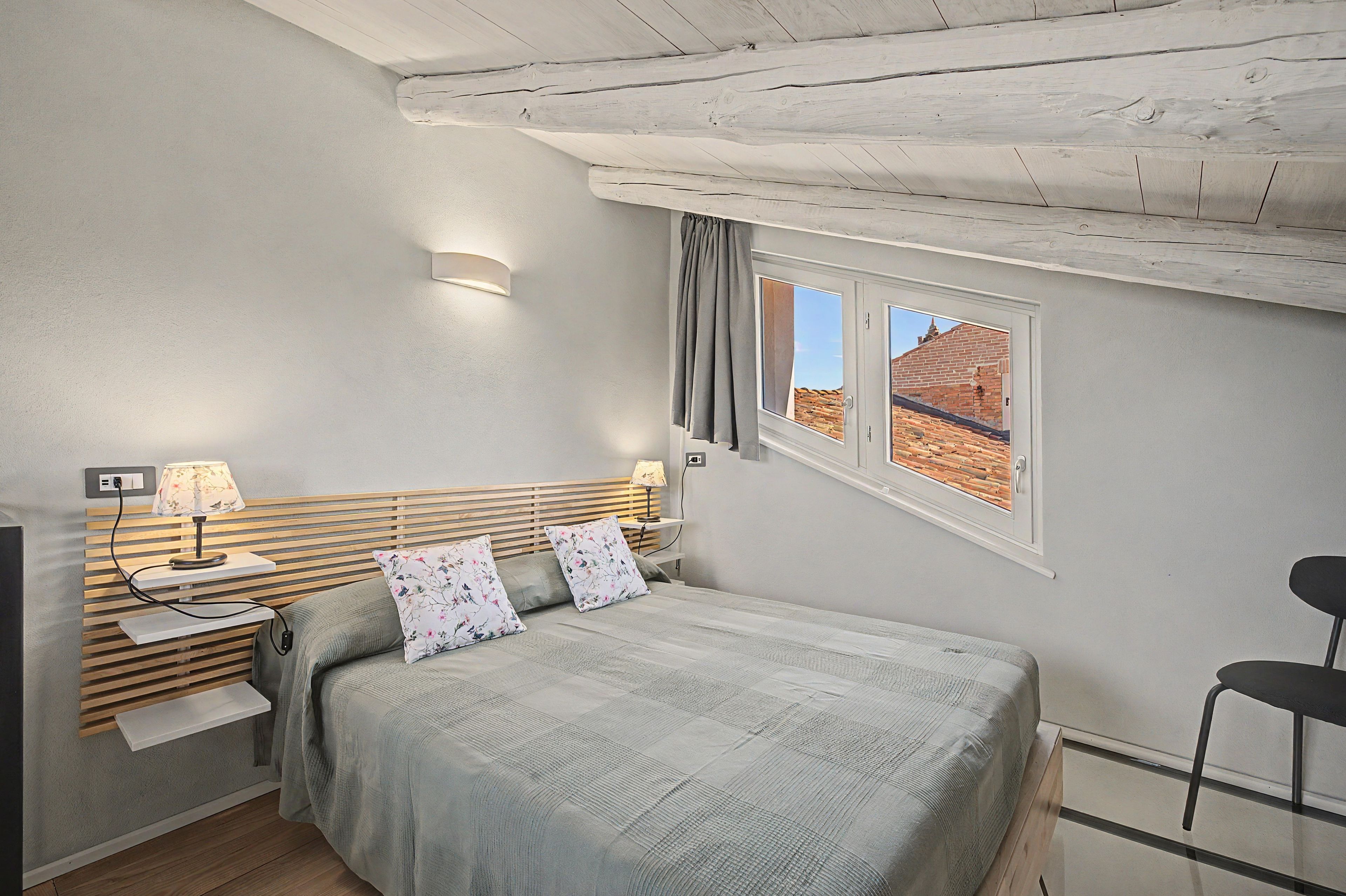 Un dormitorio con cama, mesitas de noche, lámpara, ventana con cortinas y una silla.
