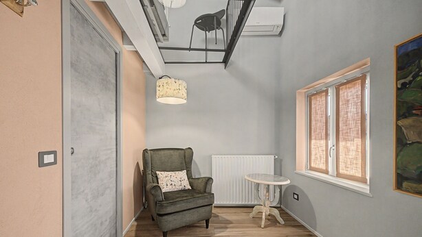 Ein Zimmer mit grauem Sessel, einem kleinen runden Tisch, einem Heizkörper und einem Fenster mit Jalousien.
