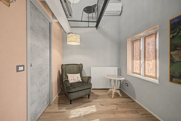 A room with a grey armchair, a small round table, a radiator, and a window with blinds.