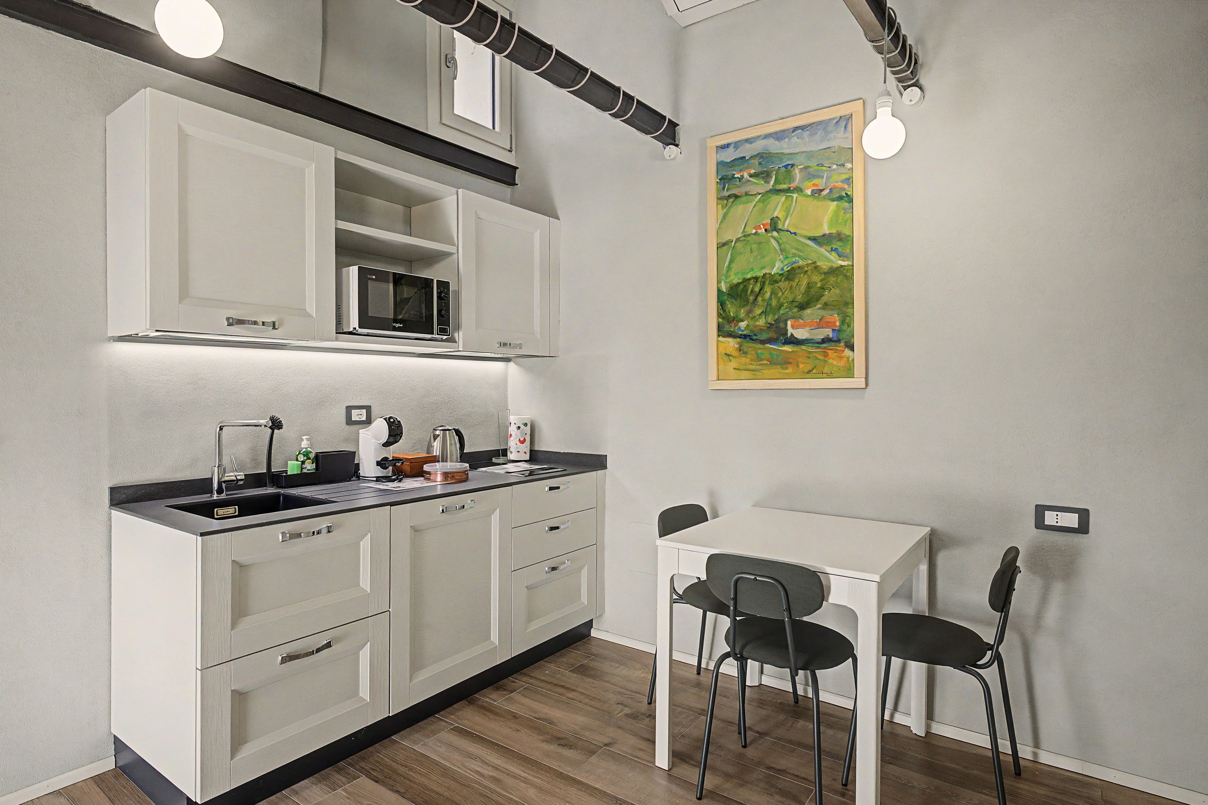 A compact kitchenette with a microwave, sink, and storage cabinets. There is a small dining table with two chairs and a painting on the wall.