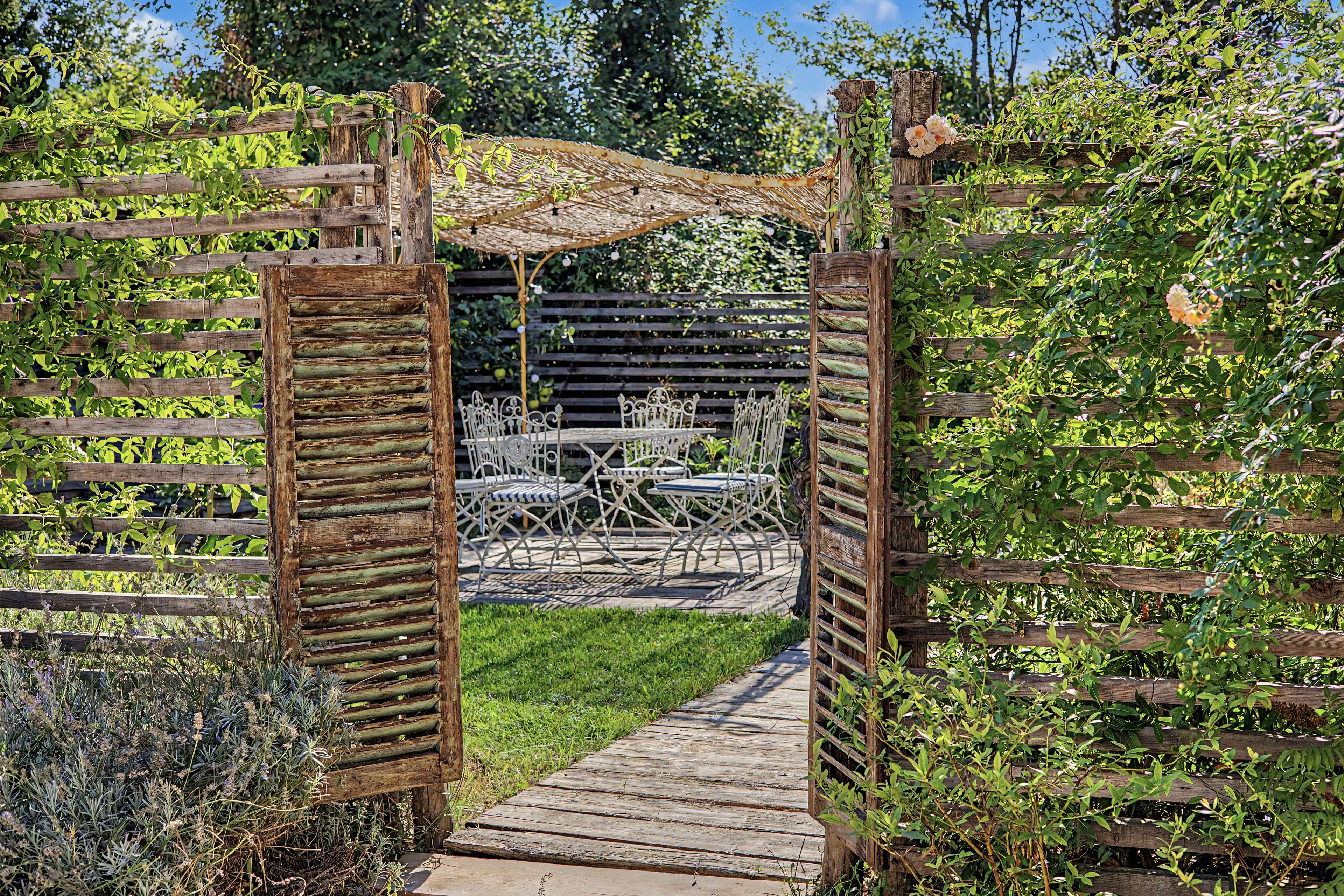 A wooden pergola with a thatched roof, a metal table and chairs, and a pathway leading through it.