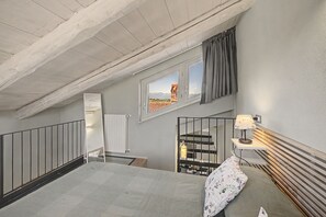 A bedroom with a sloped ceiling, a bed, a window with a view, a radiator, and a bedside table with a lamp.