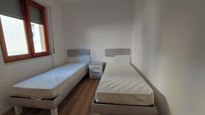 Pieni, kapea huone, jossa on kaksi erillistä sänkyä, pieni yöpöytä ja ikkuna, jossa on sälekaihtimet.