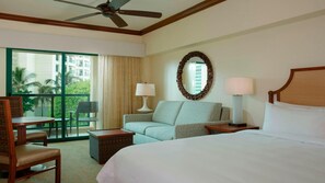 Habitación de hotel con cama, sofá, sillón, mesa, lámpara, ventilador de techo y una ventana grande con vistas a palmeras y edificios.