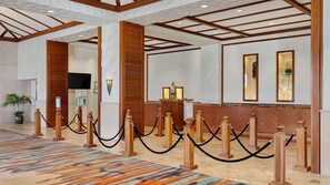 A hotel lobby with a reception desk, wooden columns, a patterned carpet, and a television screen.