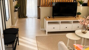 Een moderne woonkamer met een flatscreen televisie op een houten standaard, een bank, een klein rond tafeltje met een vaas, en een eethoek op de achtergrond.