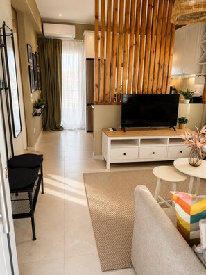 A modern living room with a flat-screen TV on a wooden stand, a sofa, a small round table with a vase, and a dining area in the background. - Nostos Home by Rentals Pro - Polychrono Halkidiki (Polychrono)