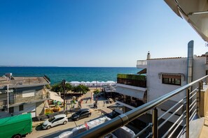 A coastal view with a clear sky, a road with parked cars, and a building with a balcony overlooking the sea.