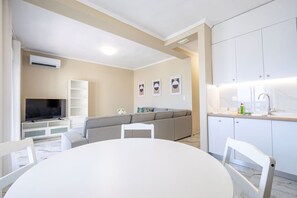 A modern living space with a flat-screen TV, a grey sofa, a dining table with chairs, and a kitchen area with a sink and cabinets. - Blue Avra Suite by Rentals Pro - Nea Moudania Halkidiki (Nea Moudania)