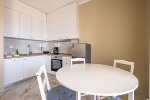 A compact kitchen with white cabinets, a built-in oven, and a round dining table with four chairs. - Blue Avra Suite by Rentals Pro - Nea Moudania Halkidiki (Nea Moudania)