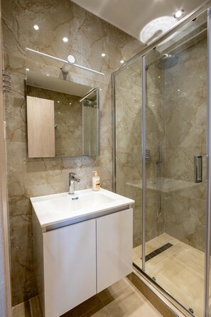A modern bathroom with a glass-enclosed shower, a white vanity with a mirror, and a marble backsplash. - Blue Avra Suite by Rentals Pro - Nea Moudania Halkidiki (Nea Moudania)