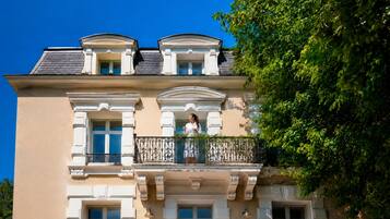 A building with a balcony, a person standing on it, and a tree to the right.