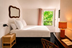 Ein Hotelzimmer mit einem Bett, einem Nachttisch mit Lampe, einem Sessel, einem Fenster mit Vorhängen und einem Spiegel an der Wand.
