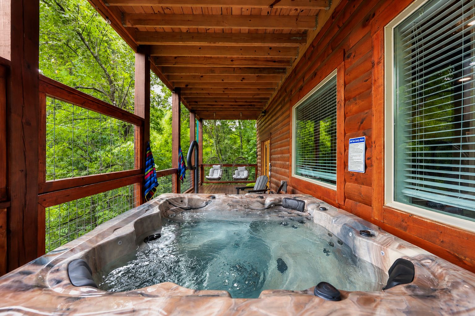 A hot tub in a wooden cabin with a covered porch, a window with blinds, and a chair outside.