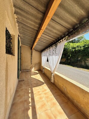 A covered balcony with a tiled floor, a wooden ceiling beam, and a view of a road and greenery.