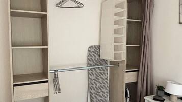 A room with a bed, a wardrobe with multiple shelves, a hanging rack, and a small table with a lamp.