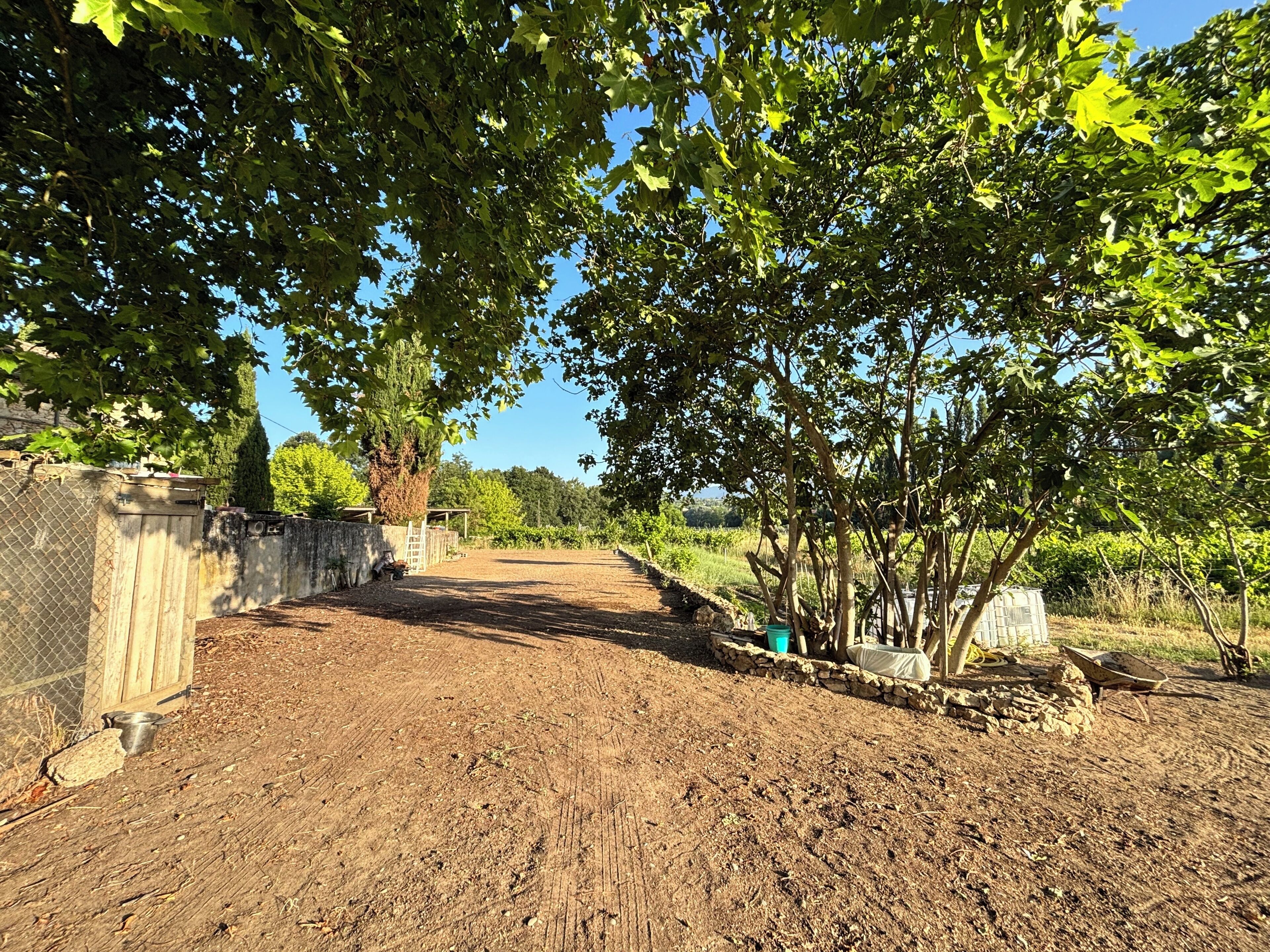 A dirt path leading to a stone wall with a metal gate, surrounded by trees and a clear sky.
