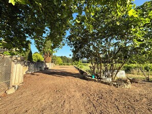 Un sendero de tierra que lleva a un muro de piedra con una verja de metal, rodeado de árboles y un cielo despejado.