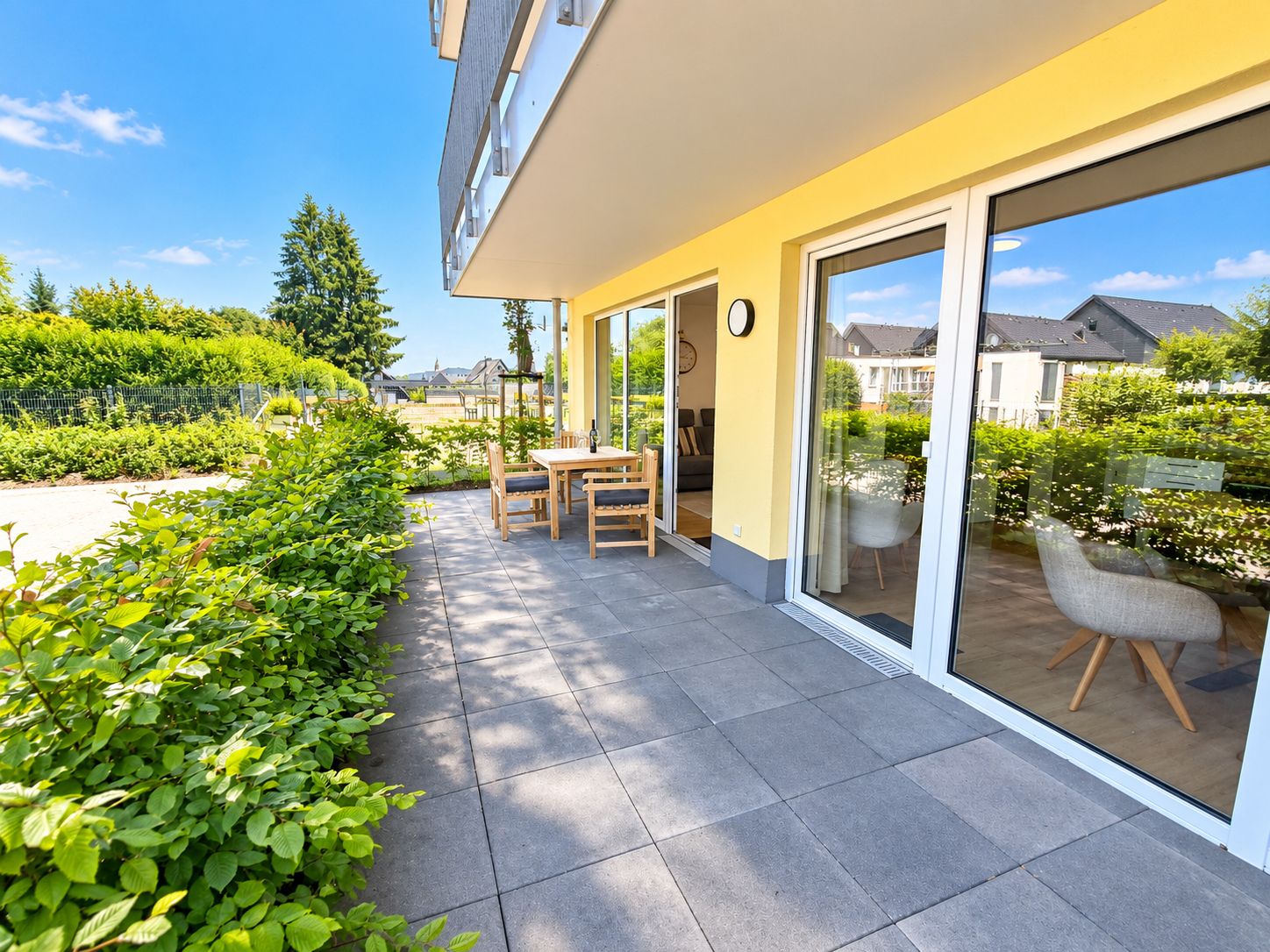A modern outdoor patio with a dining table and chairs, a glass door leading to a garden, and a clear view of the surrounding neighborhood.