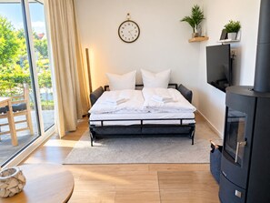 A modern bedroom with a bed, a clock, a TV, a fireplace, and a view of greenery outside. - Holiday apartment with infrared sauna, fireplace & terrace in Winterberg (Winterberg)