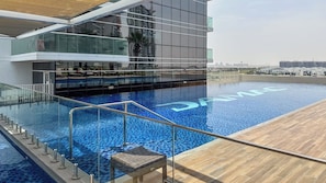 A modern rooftop pool with glass railings, a wooden deck, and a cityscape view in the background. - Maison Privee - Elegant Studio Retreat in Damac Hills (Dubai)