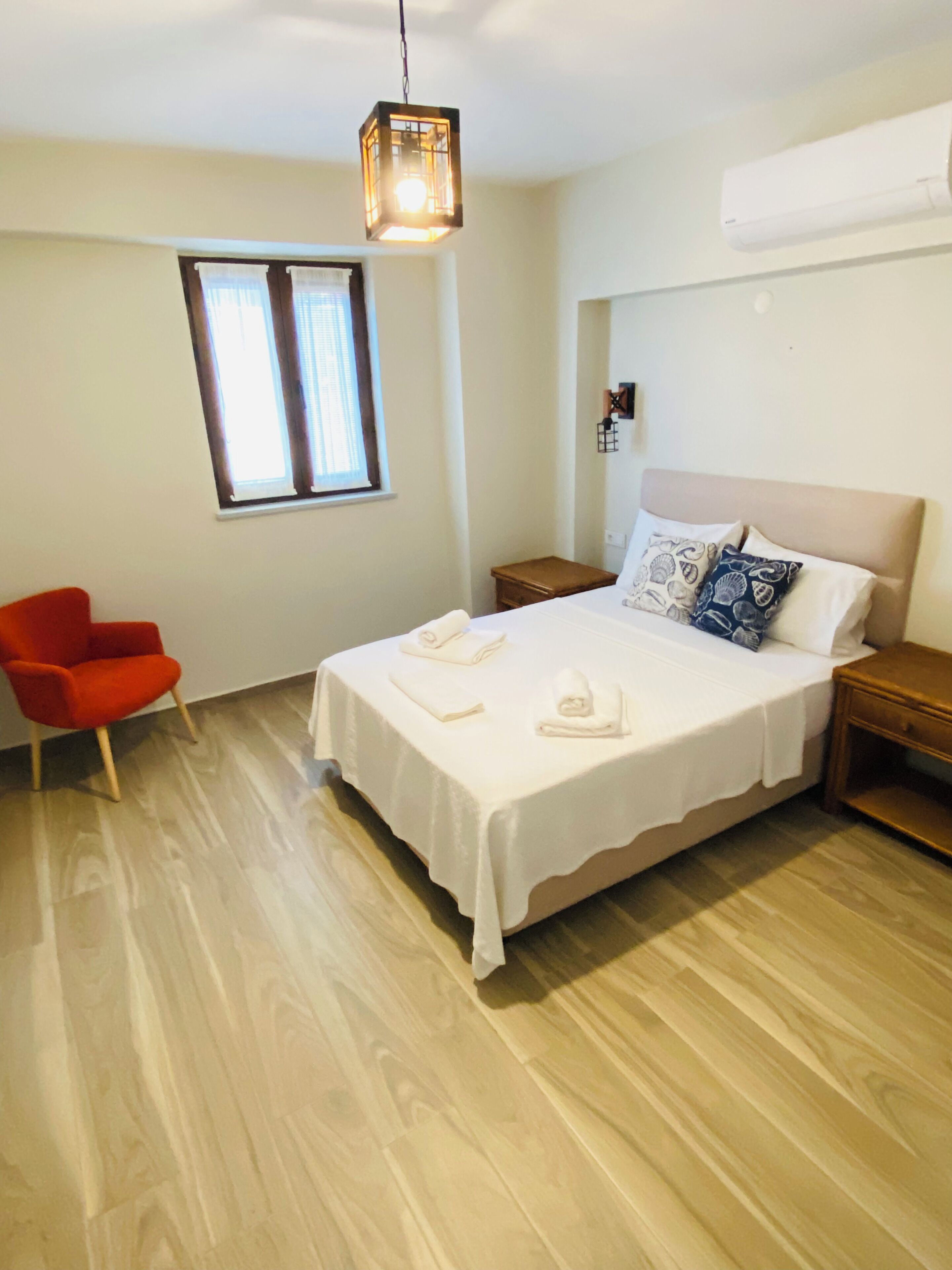 Un dormitorio con cama, mesita de noche, una silla roja, ventana y lámpara colgante.