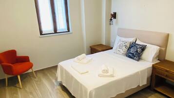 Un dormitorio con cama, mesita de noche, una silla roja, ventana y lámpara colgante.