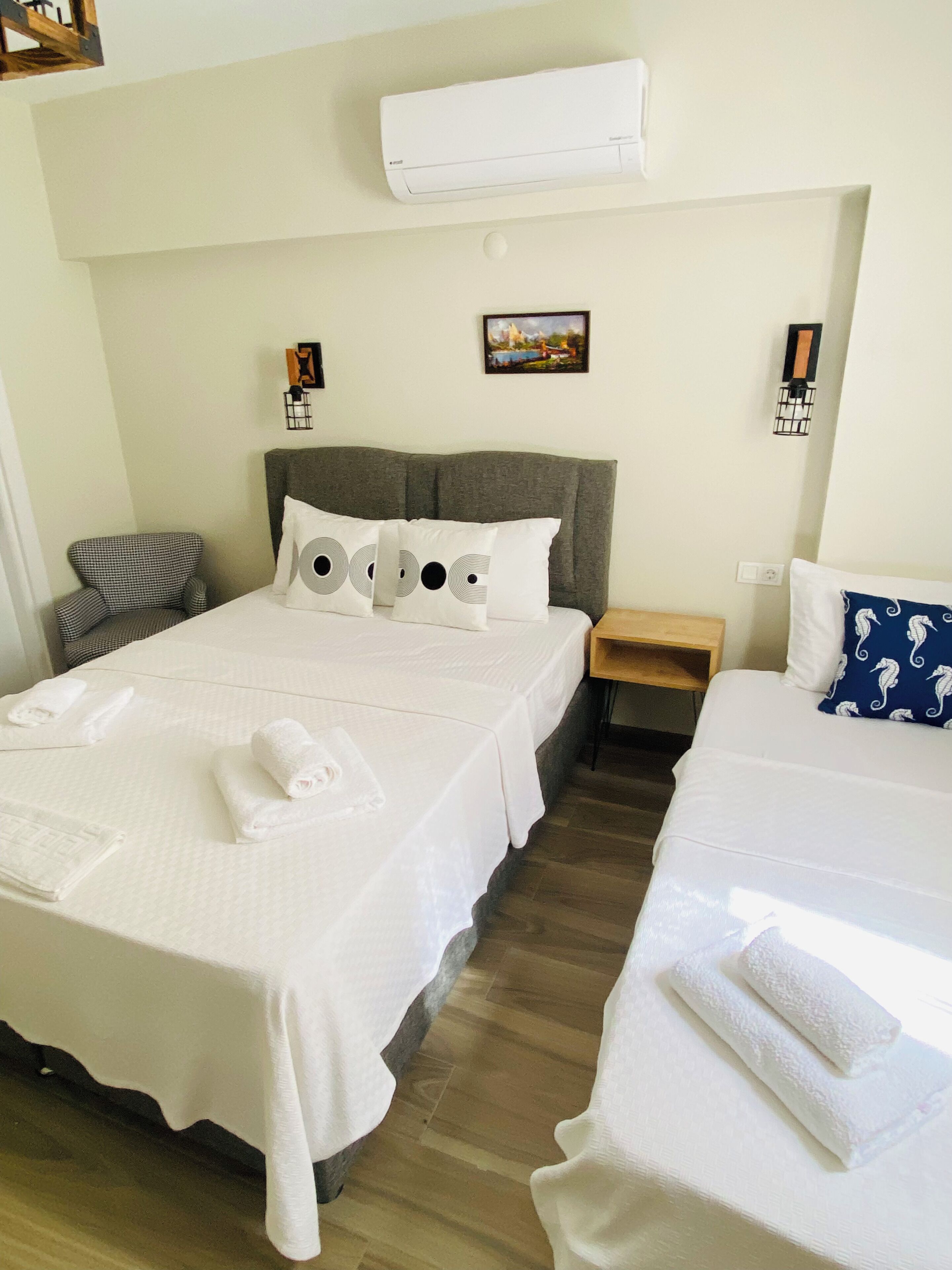 A hotel room with two beds, a gray headboard, a wall-mounted air conditioner, a small bedside table, and a chair.