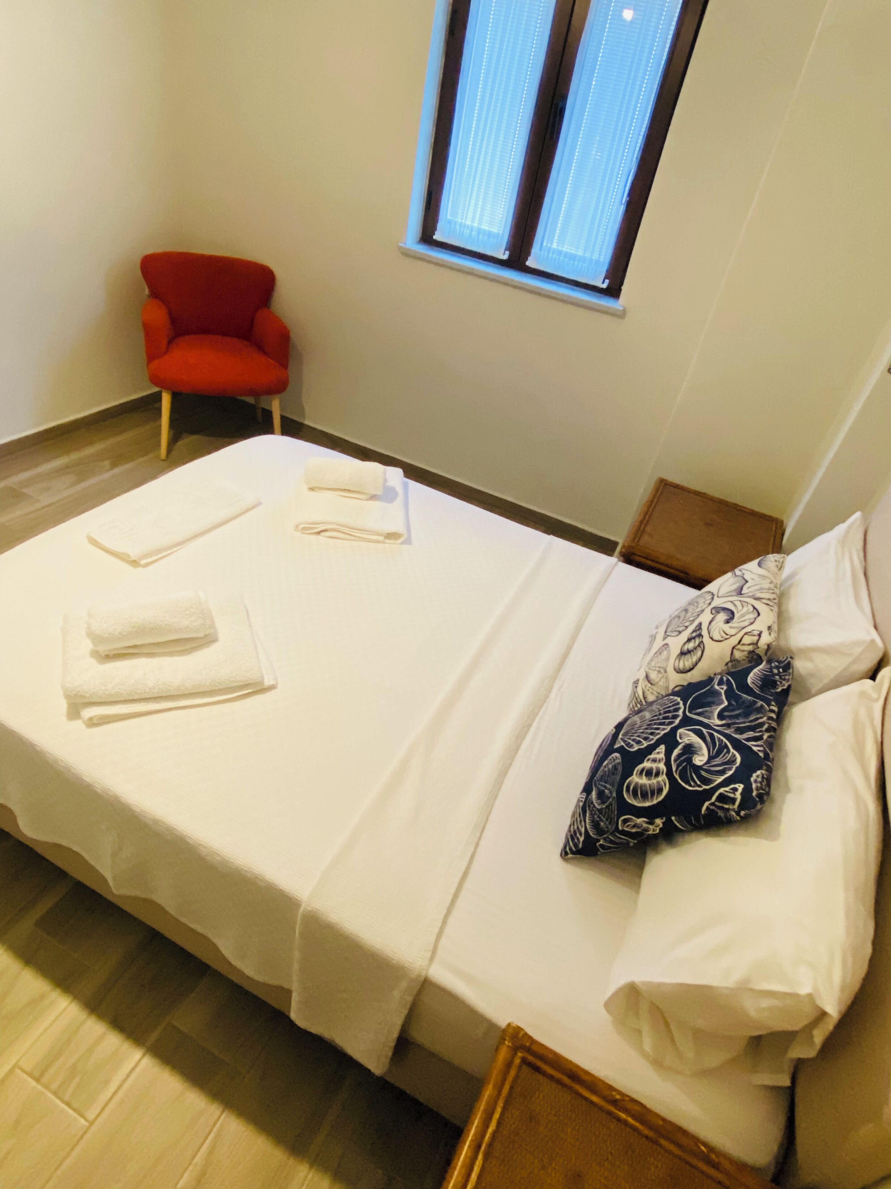 Una cama individual con ropa de cama blanca, una mesita de noche, una silla roja y una ventana con persianas.