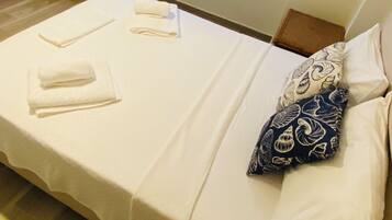 Una cama individual con ropa de cama blanca, una mesita de noche, una silla roja y una ventana con persianas.