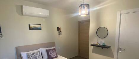 A hotel room with a bed, a bedside table, a nightstand, an air conditioner, a wall-mounted picture, a hanging light fixture, a door, and a small shelf with a mirror.