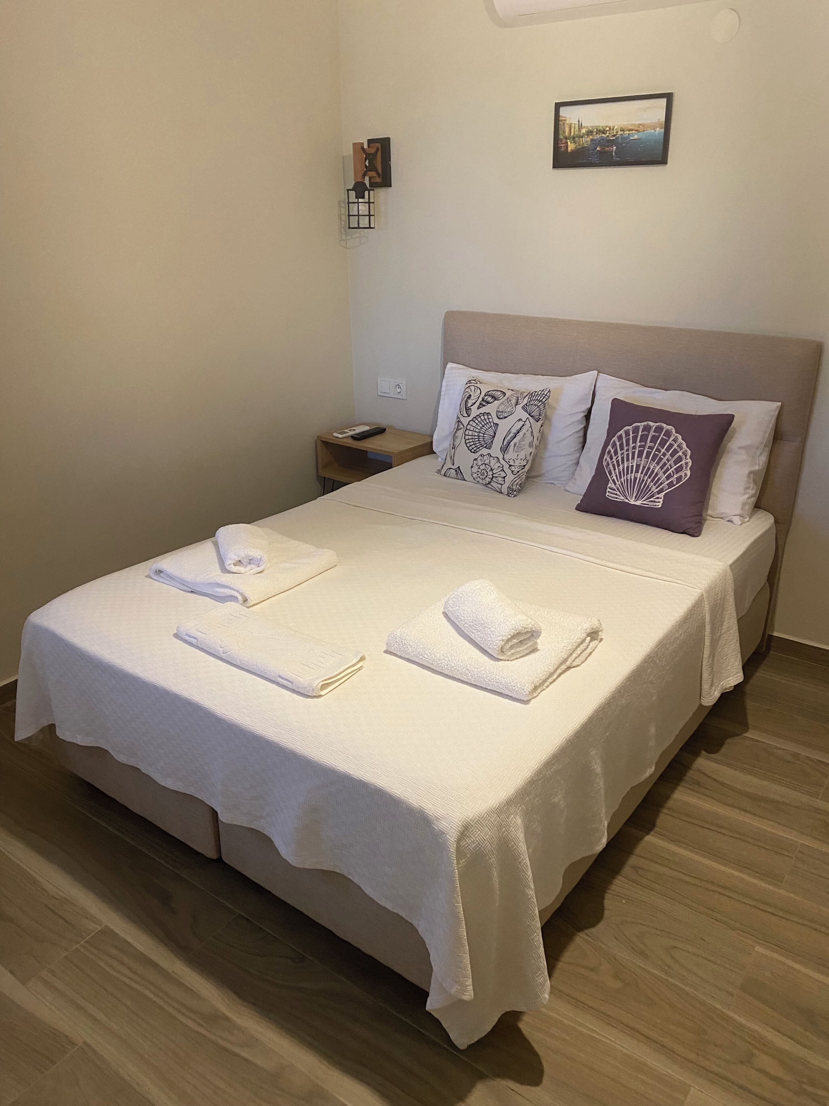Ein ordentlich bezogenes Bett mit Handtüchern, ein Nachttisch mit Fernbedienung und ein an der Wand hängendes Bild.