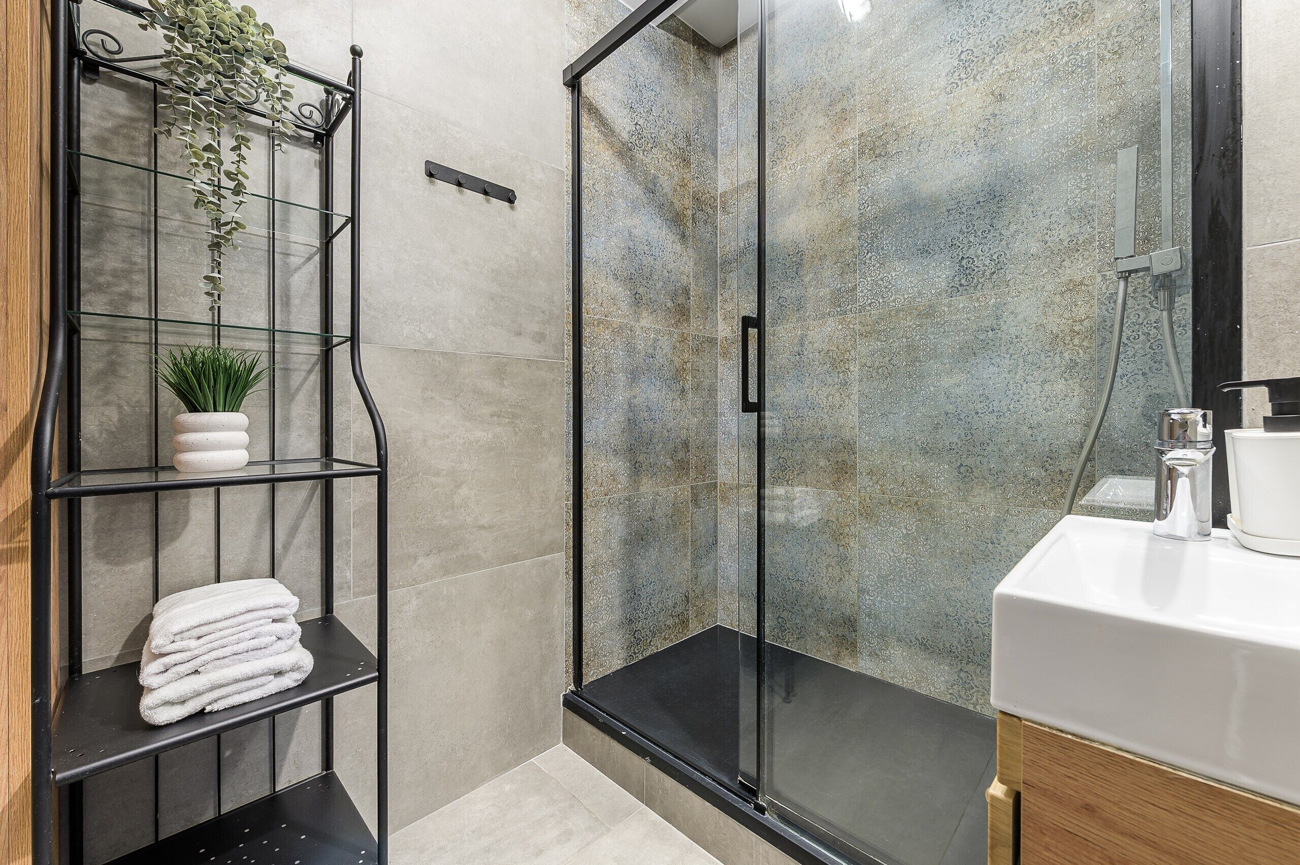 A modern bathroom with a glass shower enclosure, a white sink, and a metal shelf with plants and towels.