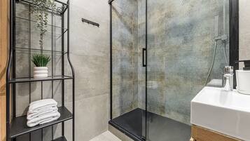 A modern bathroom with a glass shower enclosure, a white sink, and a metal shelf with plants and towels.