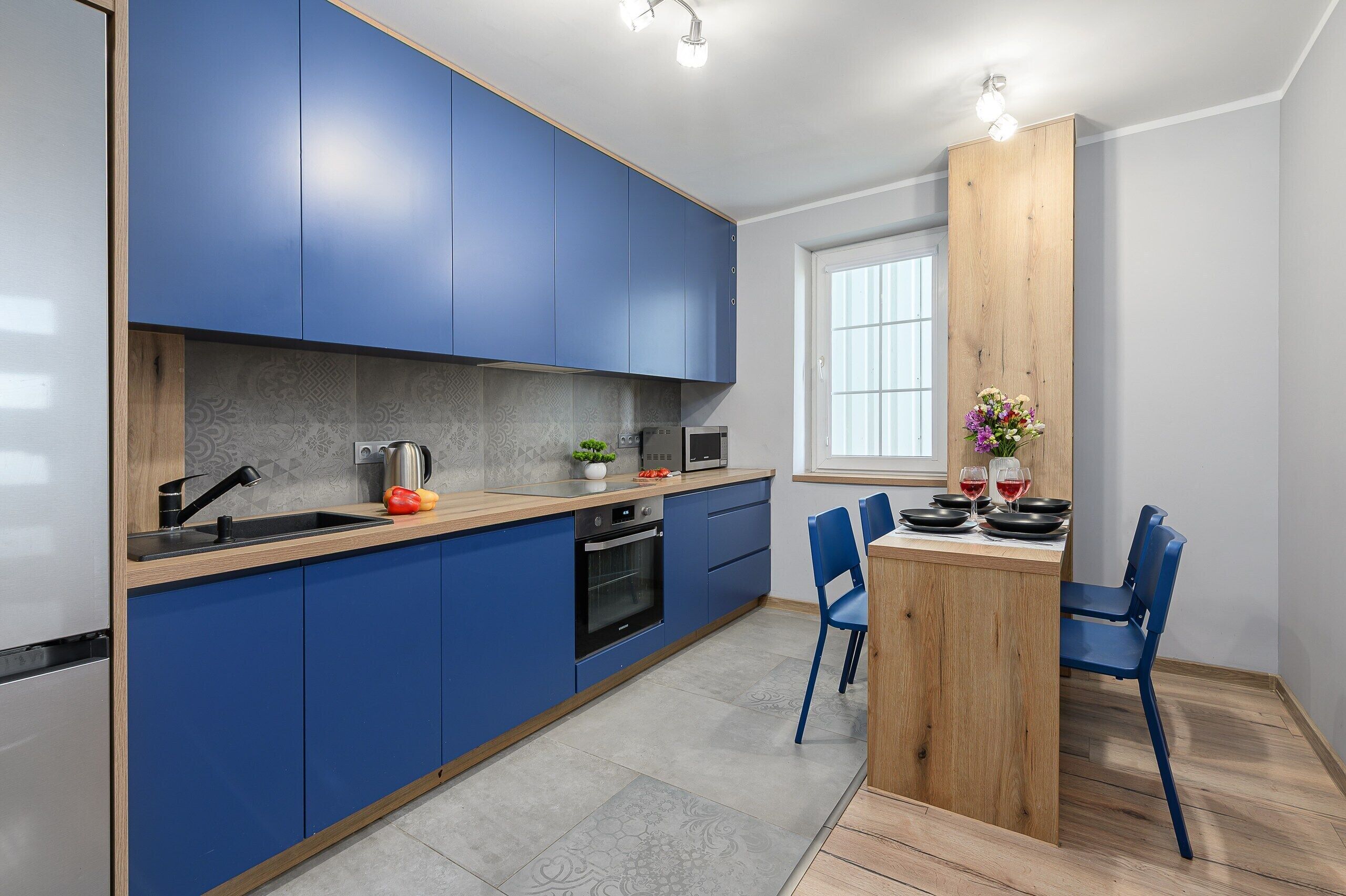 A modern kitchen with blue cabinets, a wooden dining table, and a window providing natural light.