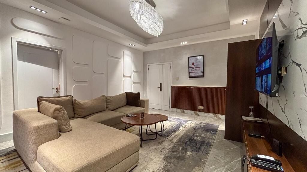 A modern living room with a sectional sofa, a coffee table, a TV mounted on the wall, and a decorative chandelier.