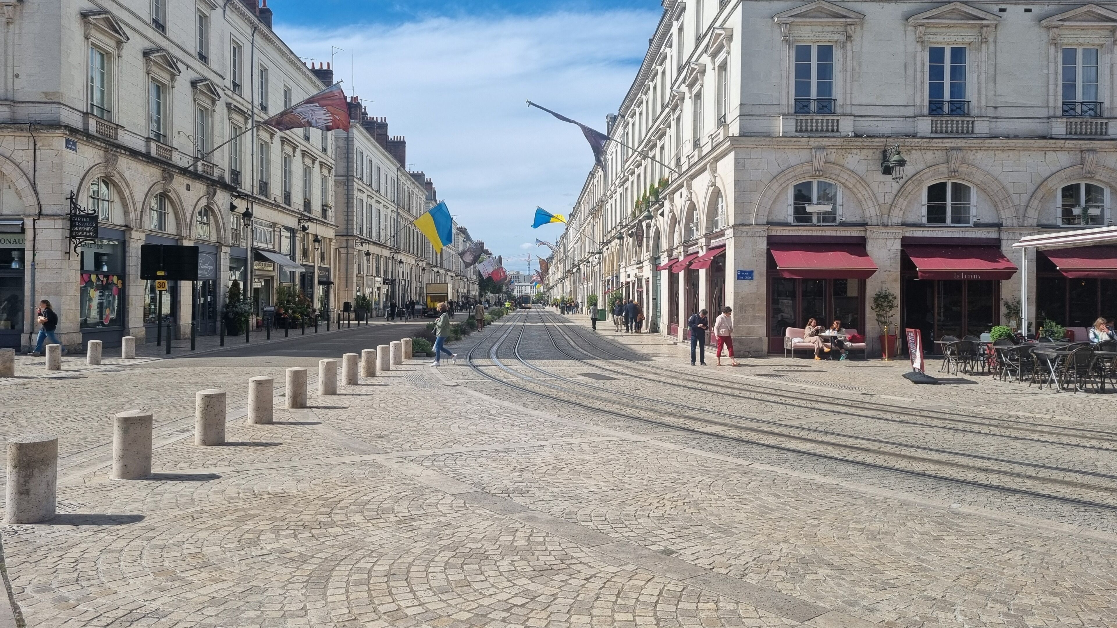A cobblestone street with tram tracks, historic buildings, and outdoor seating areas.