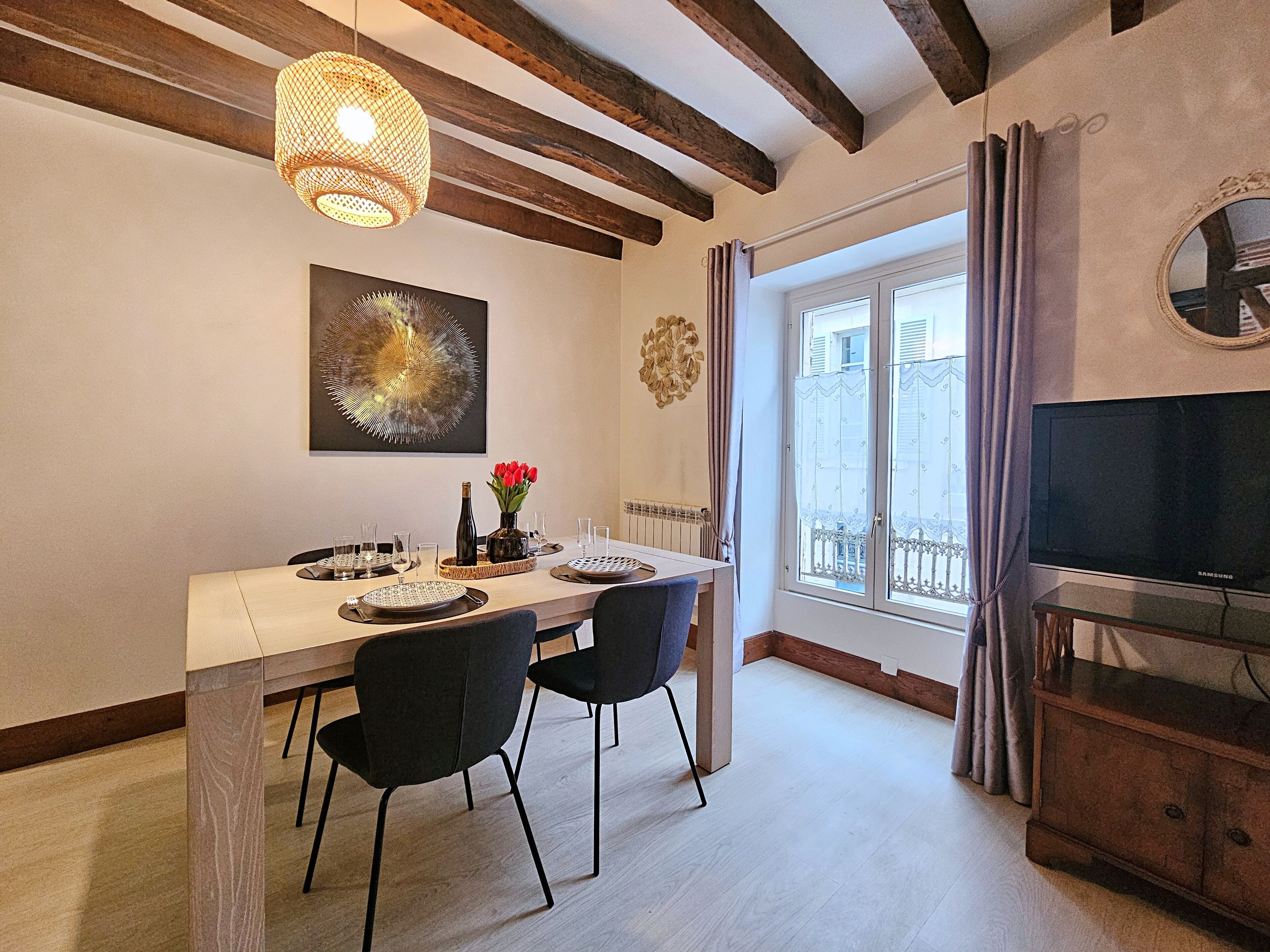 Een eetgedeelte met een tafel voor vier personen, een televisie op een standaard, en een raam met gordijnen.